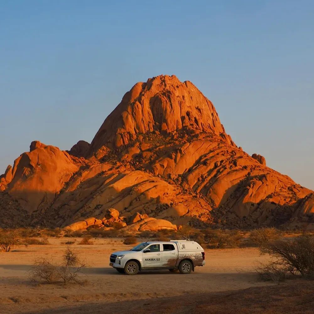 A white 4x4 safari vehicle parked in a desert landscape in front of a massive, orange, granite inselberg or mountain at sunset.