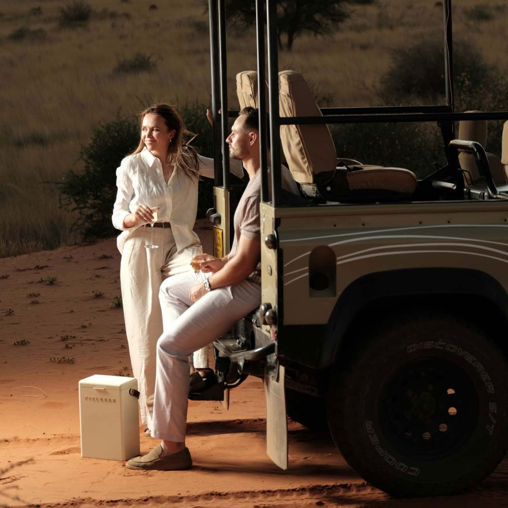 Young couple sitting near parked safari vehicle on the dunes, drinking wine.