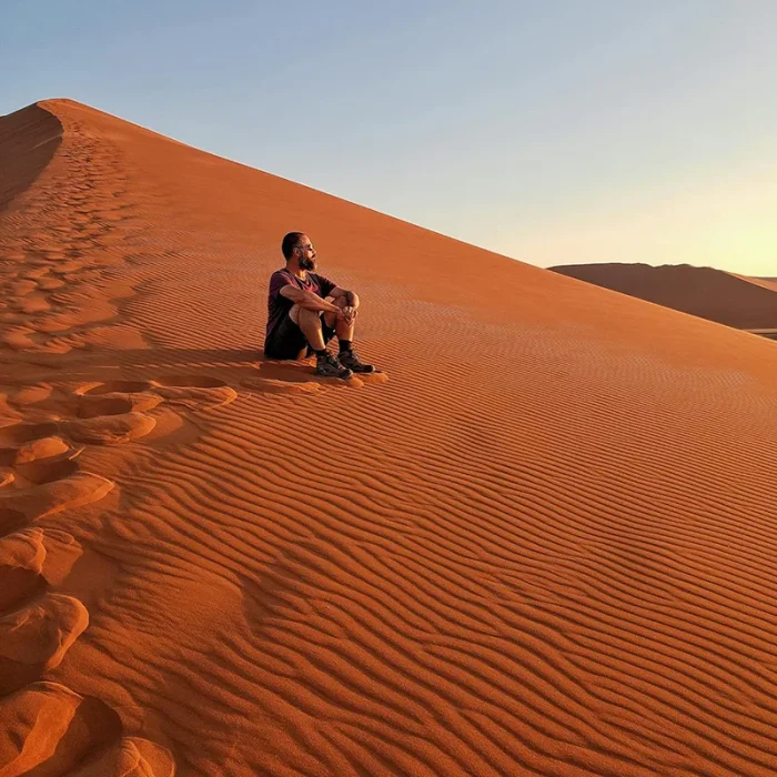 Man sitting on a ripple-textured red sand dune at sunset.